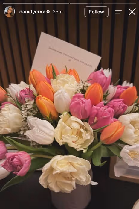 The image depicts a vibrant bouquet of multicolored flowers, including white, pink and orange tulips, and an envelope rests on top of the arrangement.