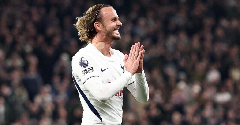 Soccer player James Maddison, wearing a white Spurs home shirt with the Nike tick logo and club cock badge on the chest, pushes his raised hands together to celebrate a goal for Premier League team Tottenham Hotspur Football Club and looks left, smiling.